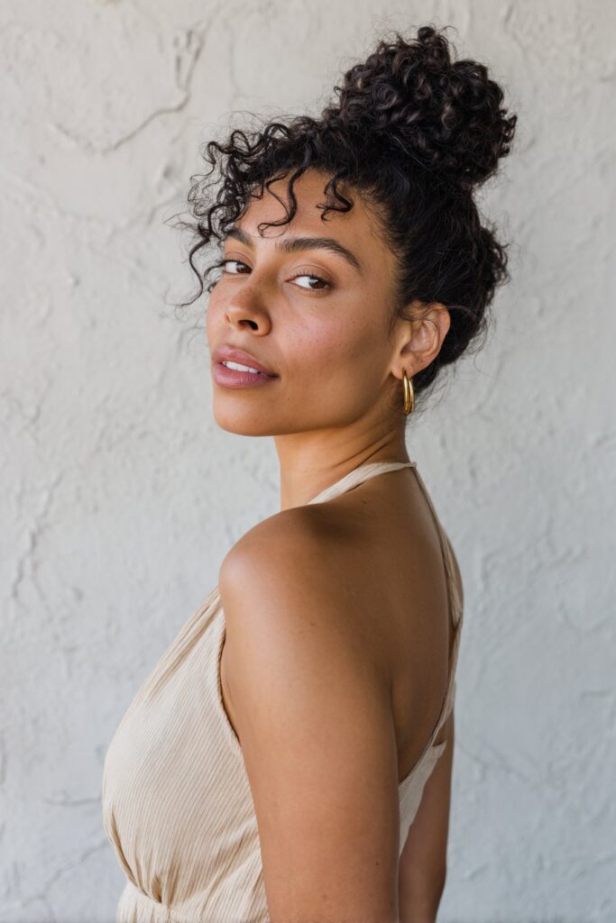 Playful curly girl updo bun for women over 60, in natural black, highlighted by a textured plaster backdrop.